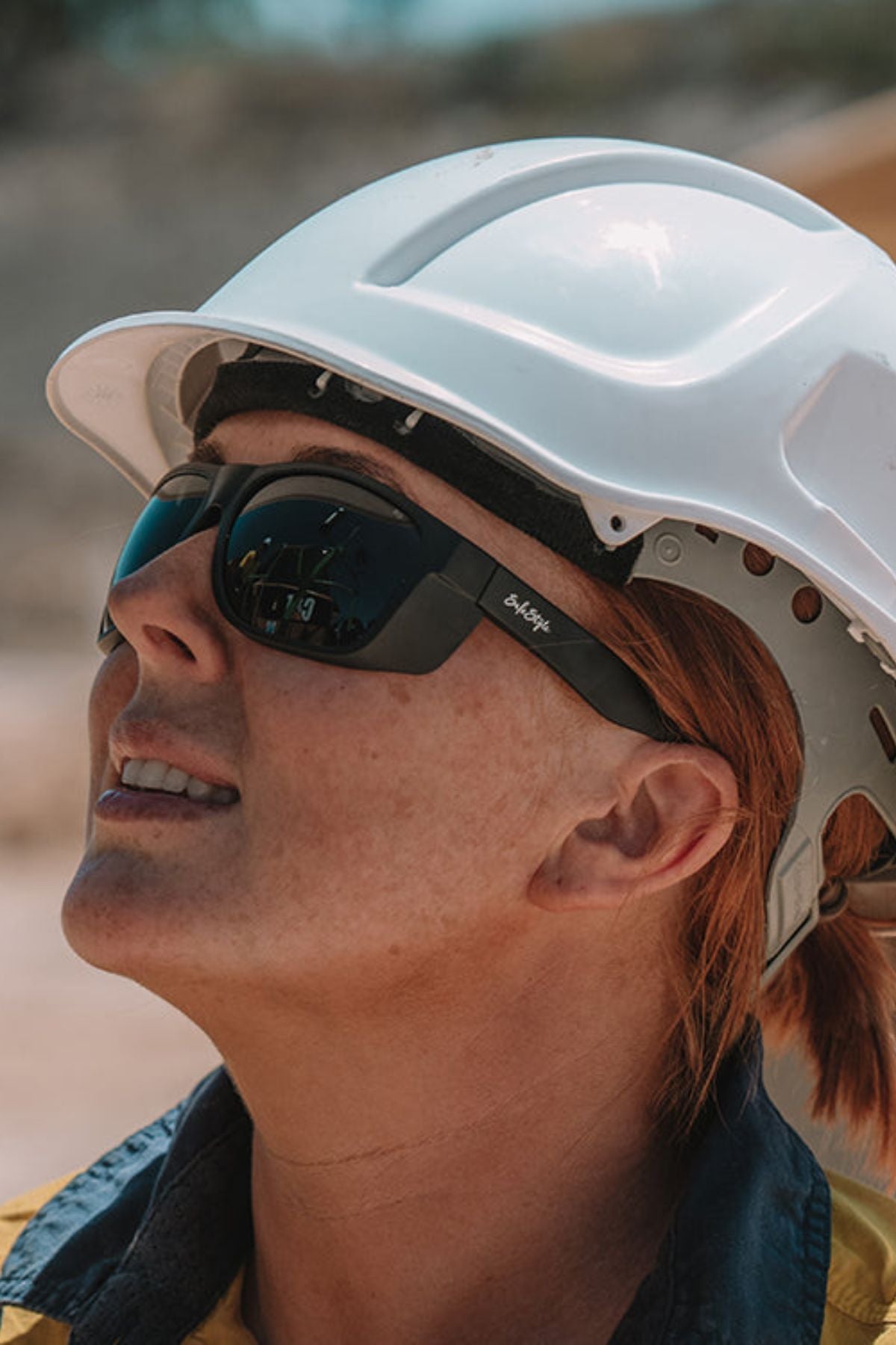 A person wearing a white hard hat, yellow safety shirt, and SafeStyle Rapids Matte Black Frame Polarised Lens sunglasses looks upward outdoors on a sunny day.