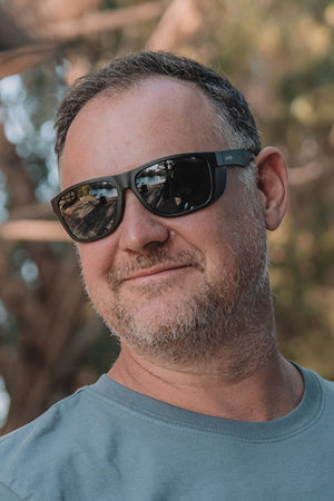 A middle-aged man with short brown hair and a light beard wears SafeStyle Rapids Matte Black Frame Polarised Lens sunglasses and a light blue T-shirt, smiling slightly outdoors with blurred trees in the background.