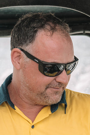 A man with short brown hair and a trimmed beard wears SafeStyle Rapids Matte Black Frame Tinted Lens sunglasses and a yellow collared shirt with a green inner collar, looking down and smiling slightly.