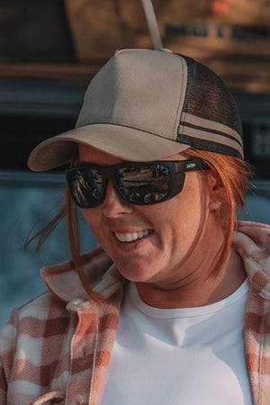 A smiling woman with red hair wears SafeStyle Rapids Matte Black Frame Tinted Lens sunglasses, a beige and black trucker hat, white shirt, and plaid jacket outdoors in casual attire.