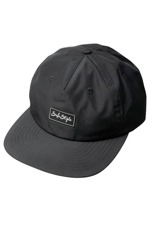 The Safestyle Worksite to Weekend Essentials Cap is a black flat-brim cap made from recycled polyester, featuring a rectangular "Safe Style" patch, stitched eyelets for ventilation, and UPF 50+ for all-day sun protection.