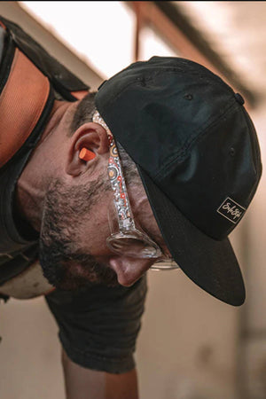 A man wearing a Safestyle Worksite to Weekend Essentials Cap, safety glasses, and orange earplugs leans forward at work. His harness strap and shirt feature quick-dry technology for comfort as he stays focused on the task.
