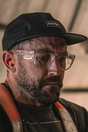 A man wears the Safestyle Worksite to Weekend Essentials Cap, made from recycled polyester, along with clear safety glasses, orange earplugs, and an orange strap, looking downward with a serious expression indoors.