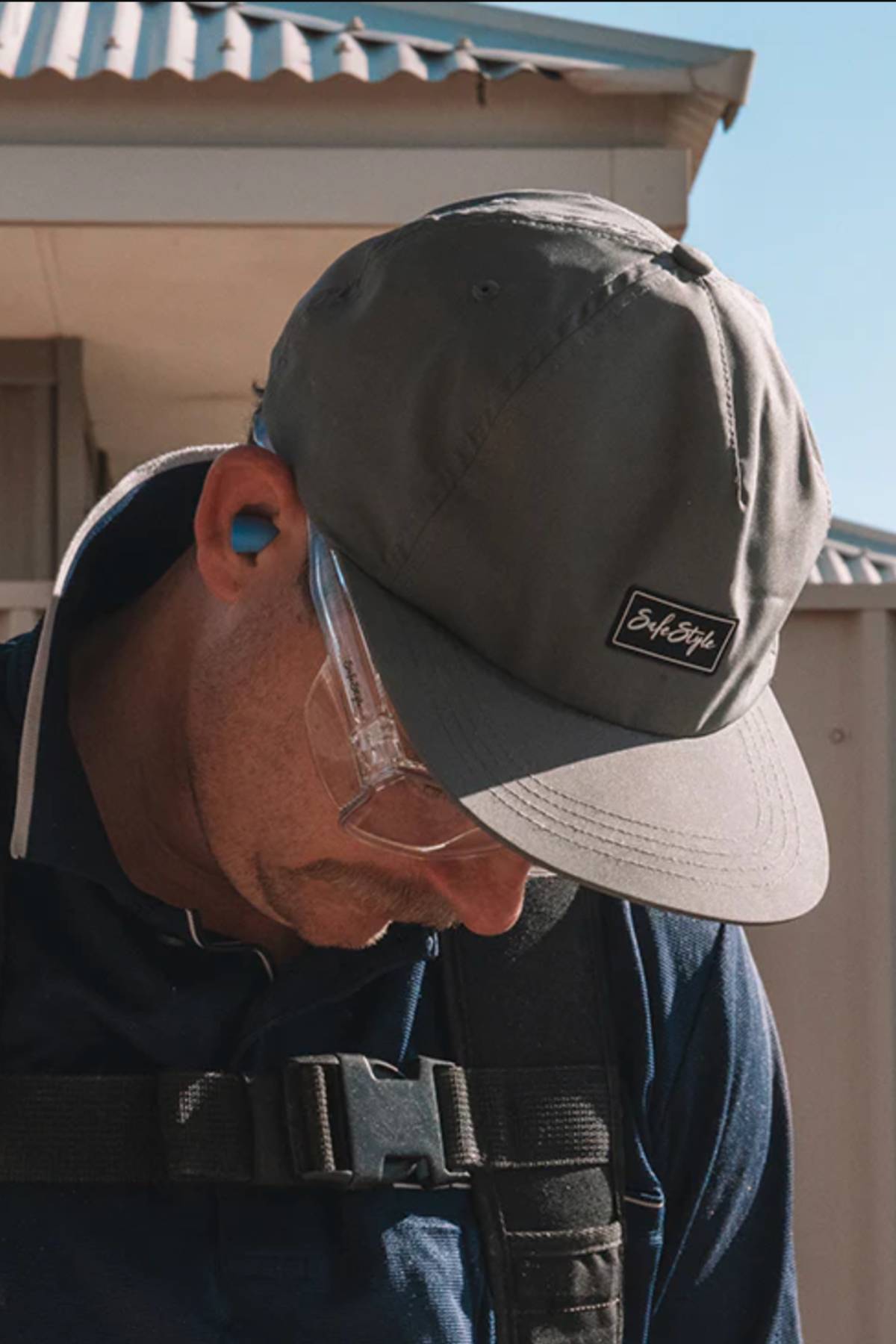 Someone wearing the Safestyle Worksite to Weekend Essentials Cap (gray, UPF 50+), clear safety glasses, blue earplugs, and a navy shirt with backpack straps looks downward outdoors on a sunny day.