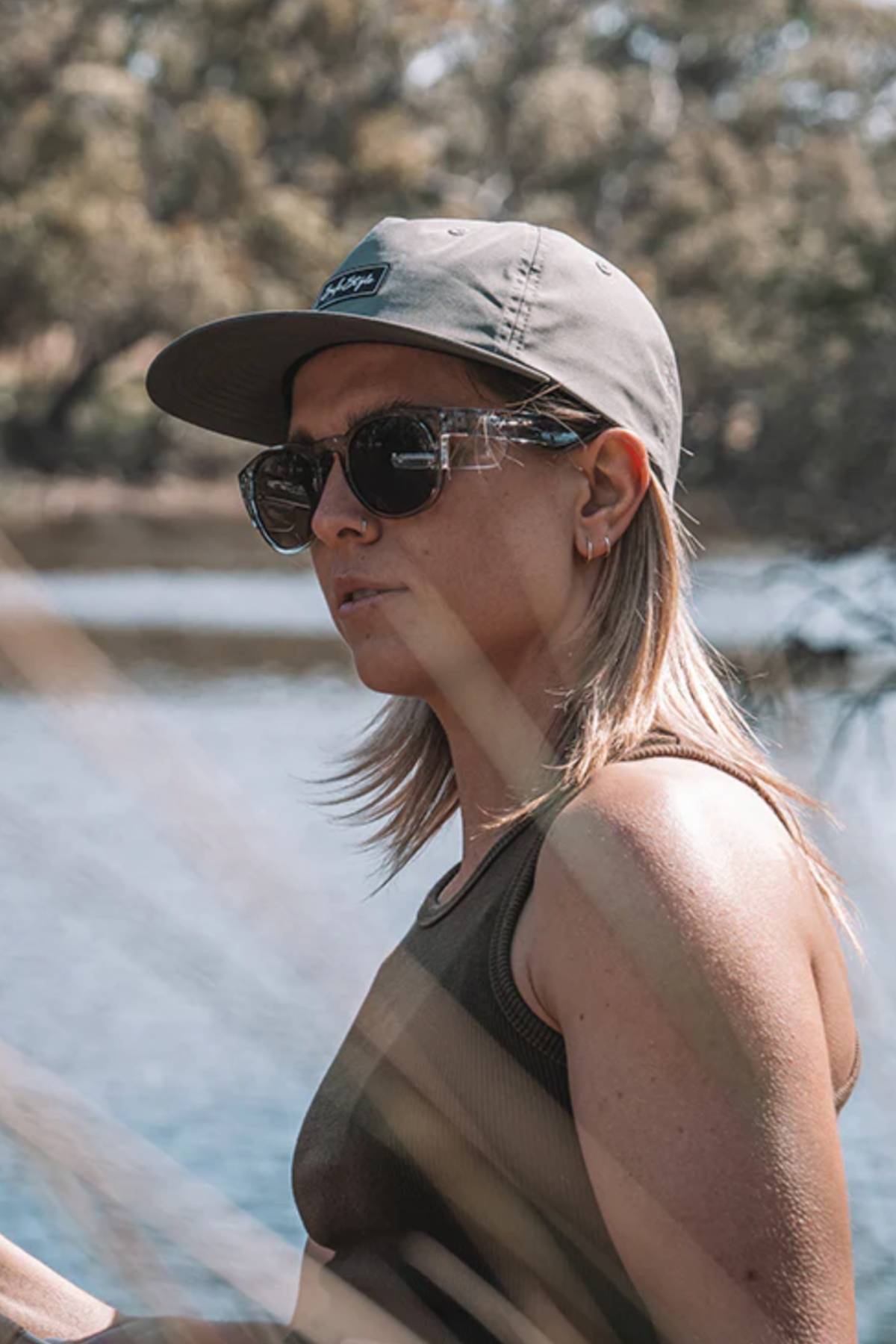 Wearing the Safestyle Worksite to Weekend Essentials Cap in gray, made from 100% recycled polyester, and black sunglasses, a person stands near water outdoors with trees behind them and sunlight casting shadows on their face.