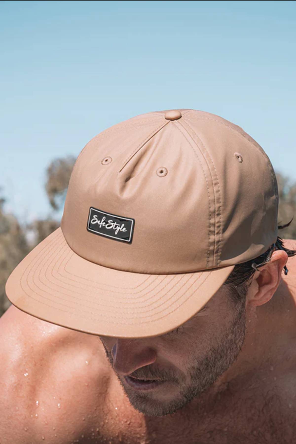 A man wears the Safestyle Worksite to Weekend Essentials Cap—a light brown recycled polyester cap with a "SafeStyle" logo and quick dry tech—outdoors against a blue sky.
