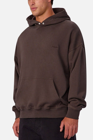 A person wearing Industrie The Sandblast Hoodie, a loose-fitting dark brown hoodie made from 100% cotton with a front pocket and hood, stands against a plain light background. The upper body shot showcases a laid-back style.