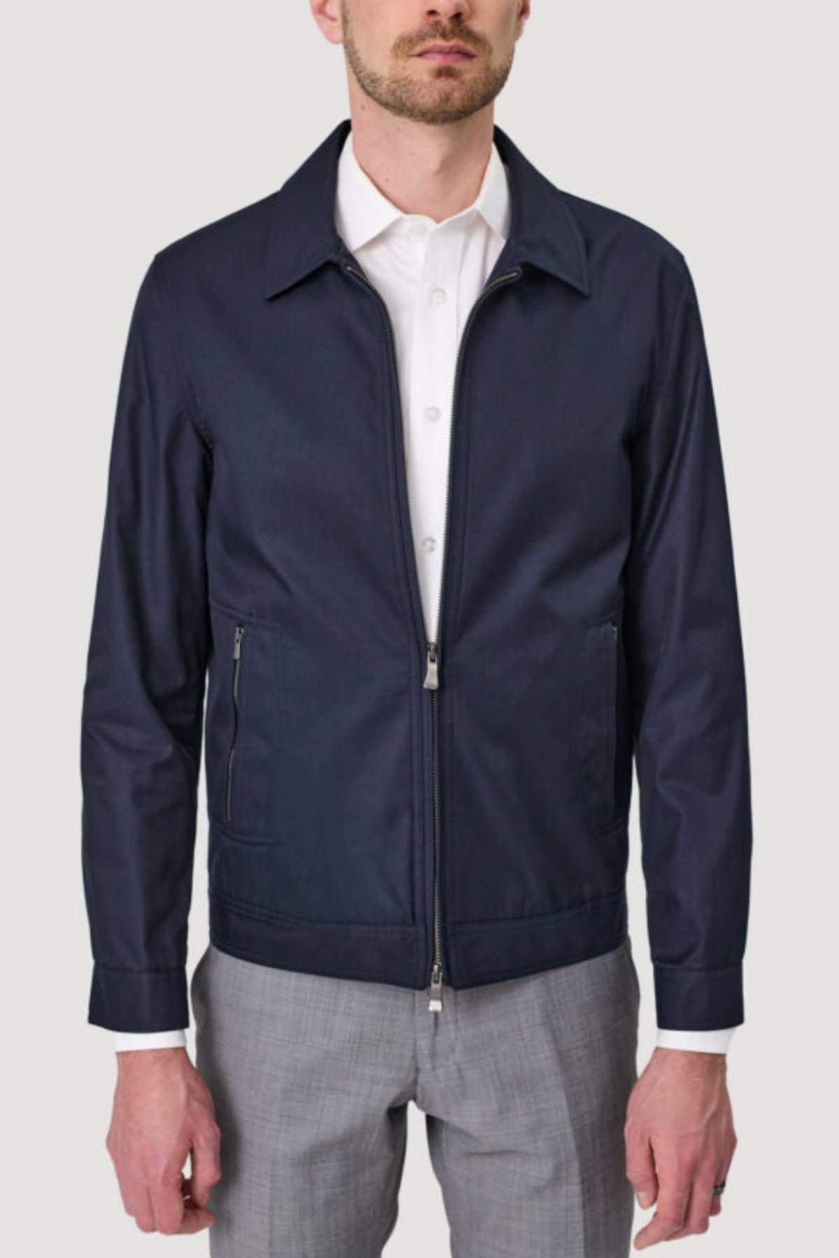 A man stands against a plain, light background, wearing the Savile Row James Zip Front Jacket in navy blue over a white dress shirt and light gray pants. His face is partially out of frame.