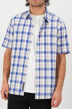 A person wears a Swanndri Coleridge Short Sleeve Shirt in blue and beige plaid over a white T-shirt, paired with black pants for a casual yet stylish look. The person's face isn't visible in the image.