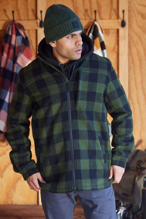 A man wearing a green and black plaid Swanndri Hudson Jacket made from New Zealand wool, gray pants, and a green knit beanie stands indoors before a wooden wall with coats and backpacks in the background.