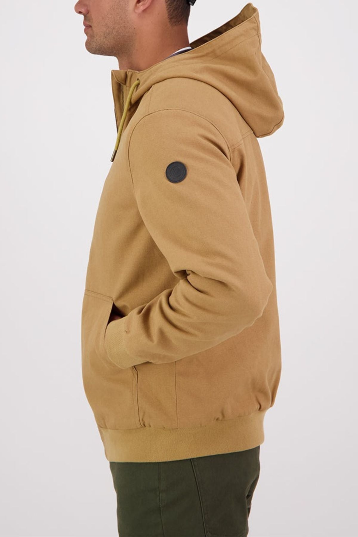 A man wearing a Swanndri Whitlow v2 Canvas Hoodieโa tan, durable 100% cotton canvas hooded sweatshirt with a black sleeve patchโand green pants stands in profile with his hands in his pockets against a plain background.