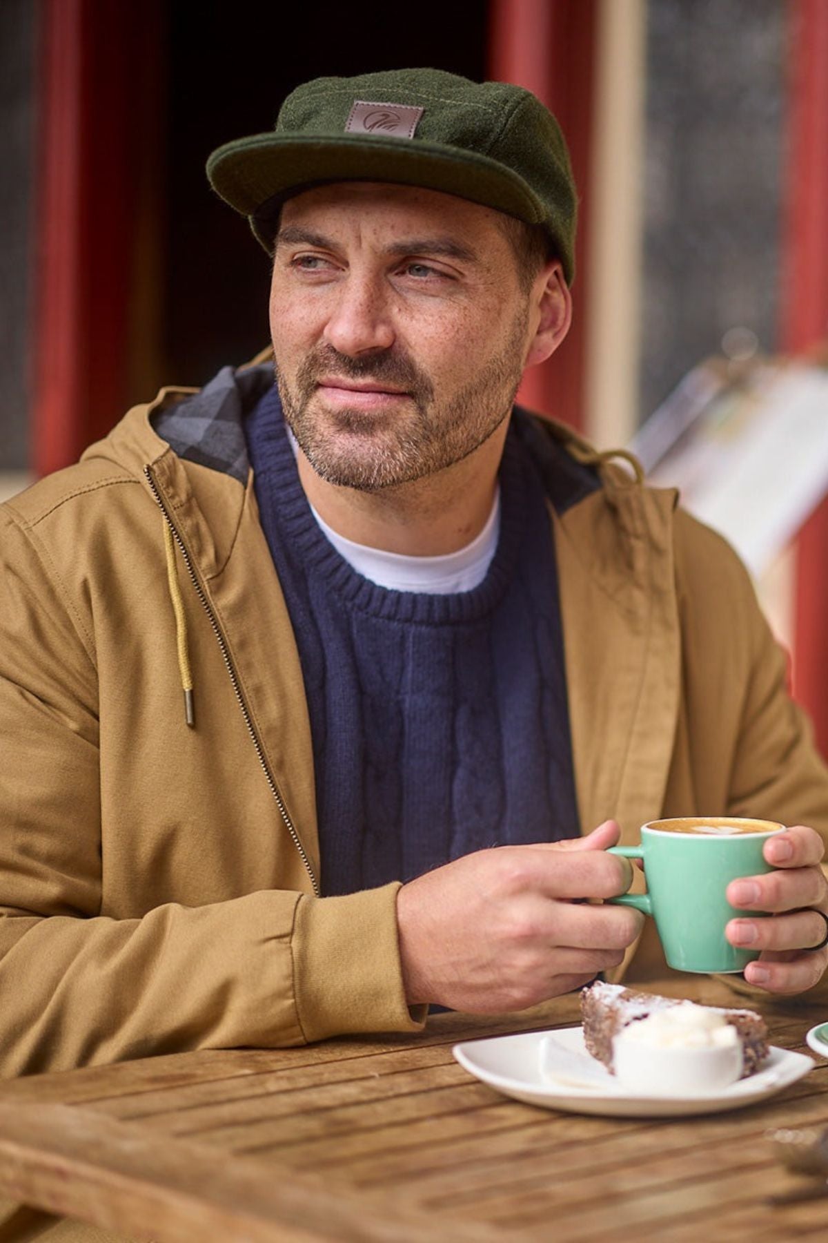 A bearded man in a green cap and a Swanndri Whitlow v2 Canvas Hoodie sits at an outdoor table, gently smiling as he holds a green coffee cup, with cake and cream on the plate before him.