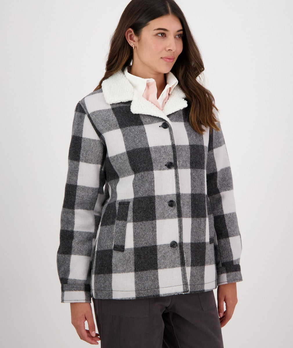 A woman with long dark hair, wearing a Swanndri Capitola Sherpa Jacket featuring a black and white checkered pattern and a white fluffy collar, stands against a plain white background. She has her hands in her pockets and is looking slightly to the right. She also wears earrings and a light pink shirt.