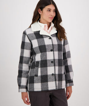A woman with long dark hair, wearing a Swanndri Capitola Sherpa Jacket featuring a black and white checkered pattern and a white fluffy collar, stands against a plain white background. She has her hands in her pockets and is looking slightly to the right. She also wears earrings and a light pink shirt.