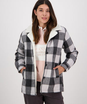 A woman is standing against a plain background wearing the Swanndri Capitola Sherpa Jacket, which features a black, white, and grey checkered pattern and a soft, white fuzzy lining. She has her hands in the jacket pockets. She has long, dark hair and is looking directly at the camera with a neutral expression.