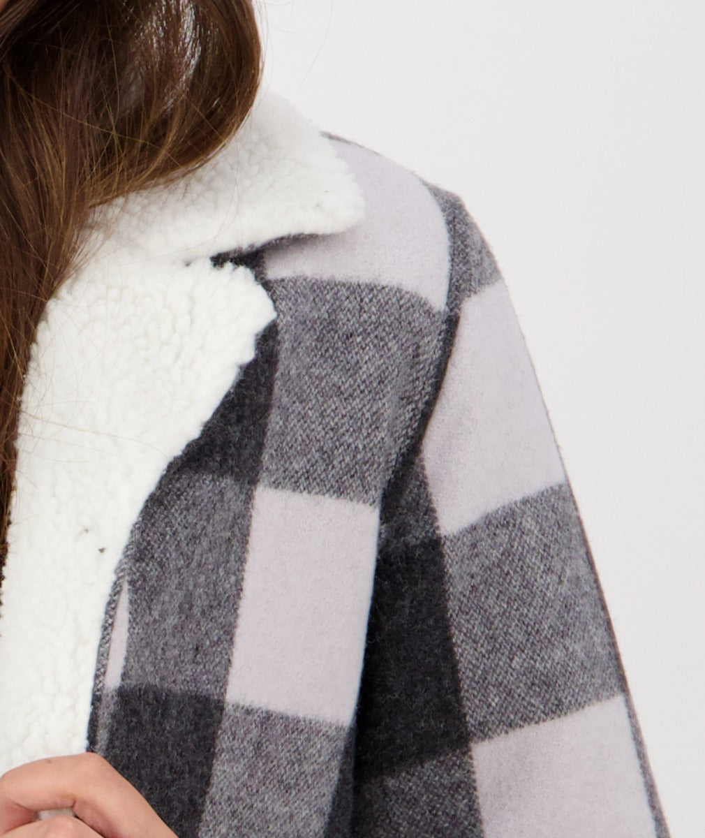 A close-up shot of a person wearing a Swanndri Capitola Sherpa Jacket featuring a gray and white checkered pattern with a thick white fleece collar. The person's hand is partially visible, holding the edge of the coat. The background is a plain white wall.