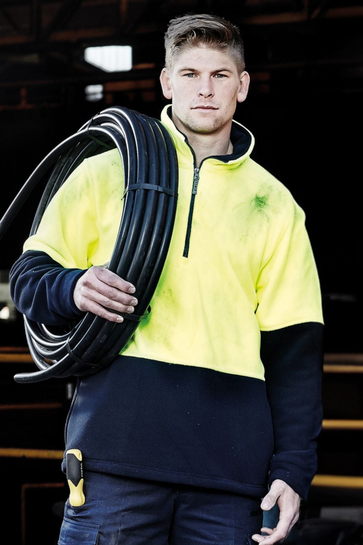 A man wearing a Syzmik Hi Vis Half Zip Polar Fleece Jumper in yellow and black holds a large coil of black cable over his shoulder, with a tool clipped to his trousers. The dimly lit background emphasizes his attire.