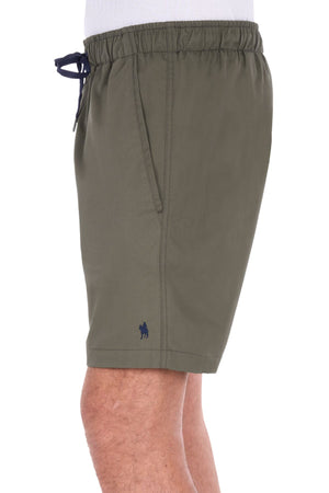 A person is seen from the side wearing olive green Thomas Cook Darcy Shorts, which have an elasticated waist with a drawstring, side pockets, and a small horse logo above the hem. These shorts are made from 100% cotton.