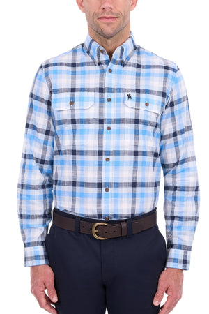 A man is wearing the Thomas Cook Jacob Shirt, a blue and white plaid button-down with chest pockets and a small embroidered logo on the left chest. The shirt is made from 100% cotton, and he has paired it with dark pants. His face is partially visible.