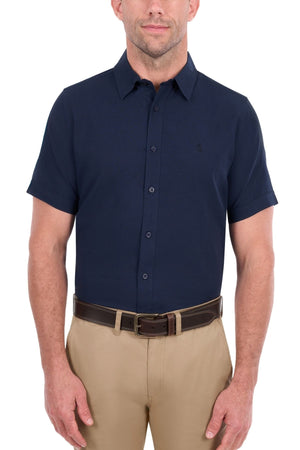 A man stands against a plain white background wearing a navy Thomas Cook Linen Short Sleeve Shirt, tucked into tan pants with a brown belt.