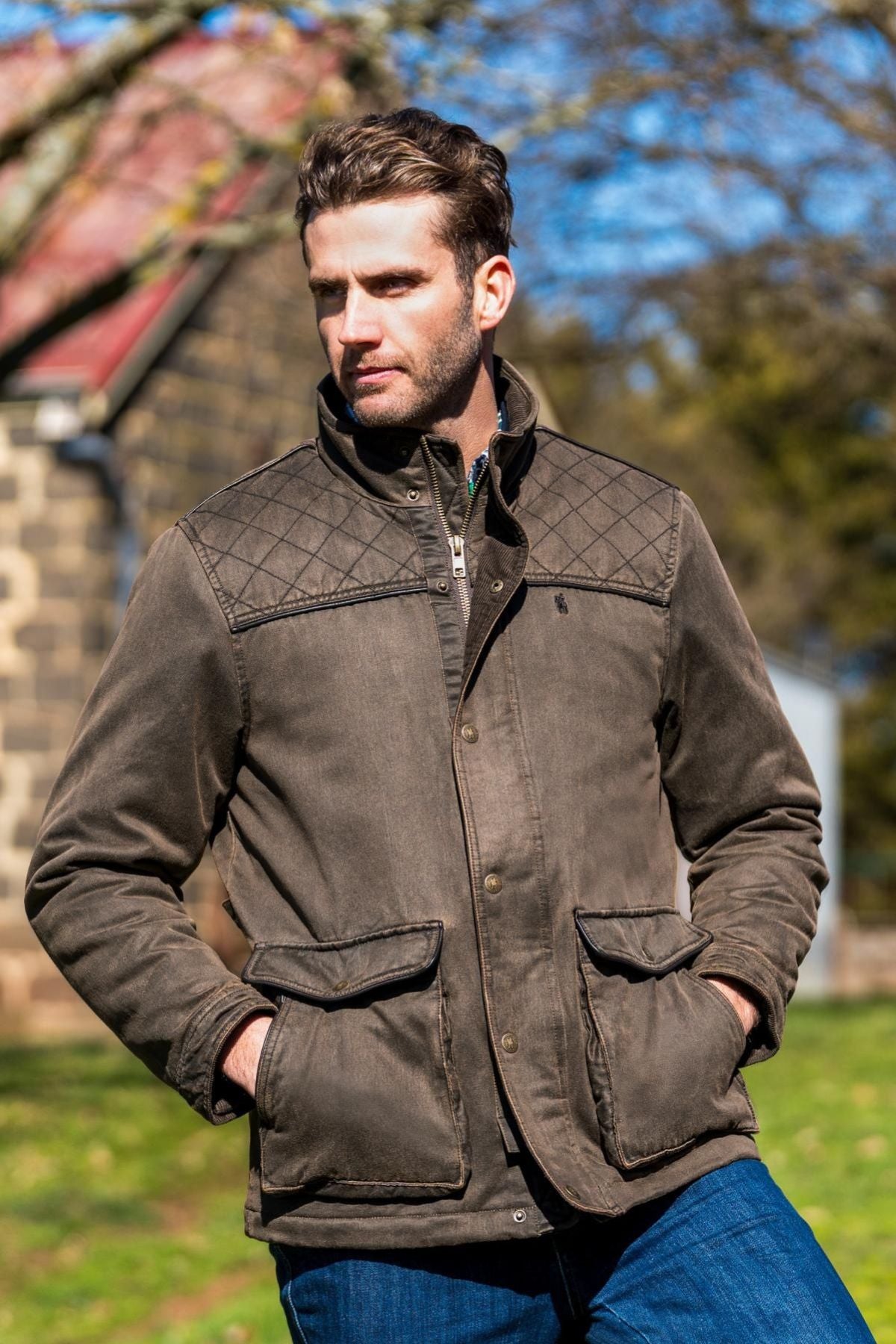 A man stands outdoors with his hands in his pockets, wearing a brown Thomas Cook Marcus Jacket with quilted shoulders and sherpa lining, paired with blue jeans. The jacket stands out against a stone building and trees in the sunshine.