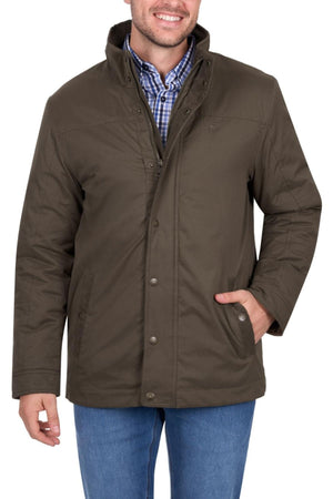 A man in a brown Thomas Cook Matt Jacket, designed for weather protection, layered over a blue and white checkered shirt with blue jeans, stands smiling with one hand in his pocket.