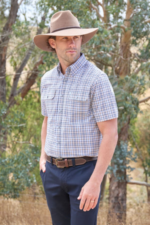 A man stands outdoors in front of trees, looking off to the side, wearing a wide-brimmed hat, dark pants, and the Thomas Cook Ted Short Sleeve Shirt.