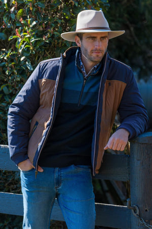 A man in a tan wide-brimmed hat, dark sweater, blue jeans, and the Thomas Cook Toby Reversible Jacket leans casually against a wooden fence with greenery behind him.