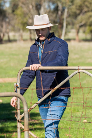 A man wearing a wide-brimmed hat, jeans, and a Thomas Cook Toby Reversible Jacket stands outdoors, opening a metal farm gate in a grassy field with trees behind him.