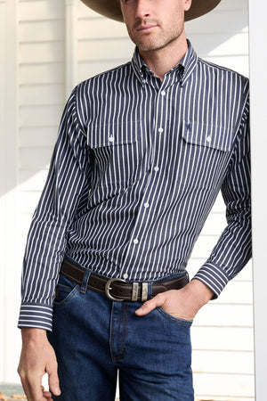 A man wearing a Thomas Cook Winton Shirt—a striped cotton shirt with double chest pockets—plus a wide-brimmed hat, dark belt, and blue jeans stands leaning against a white wall, his head partially out of frame.