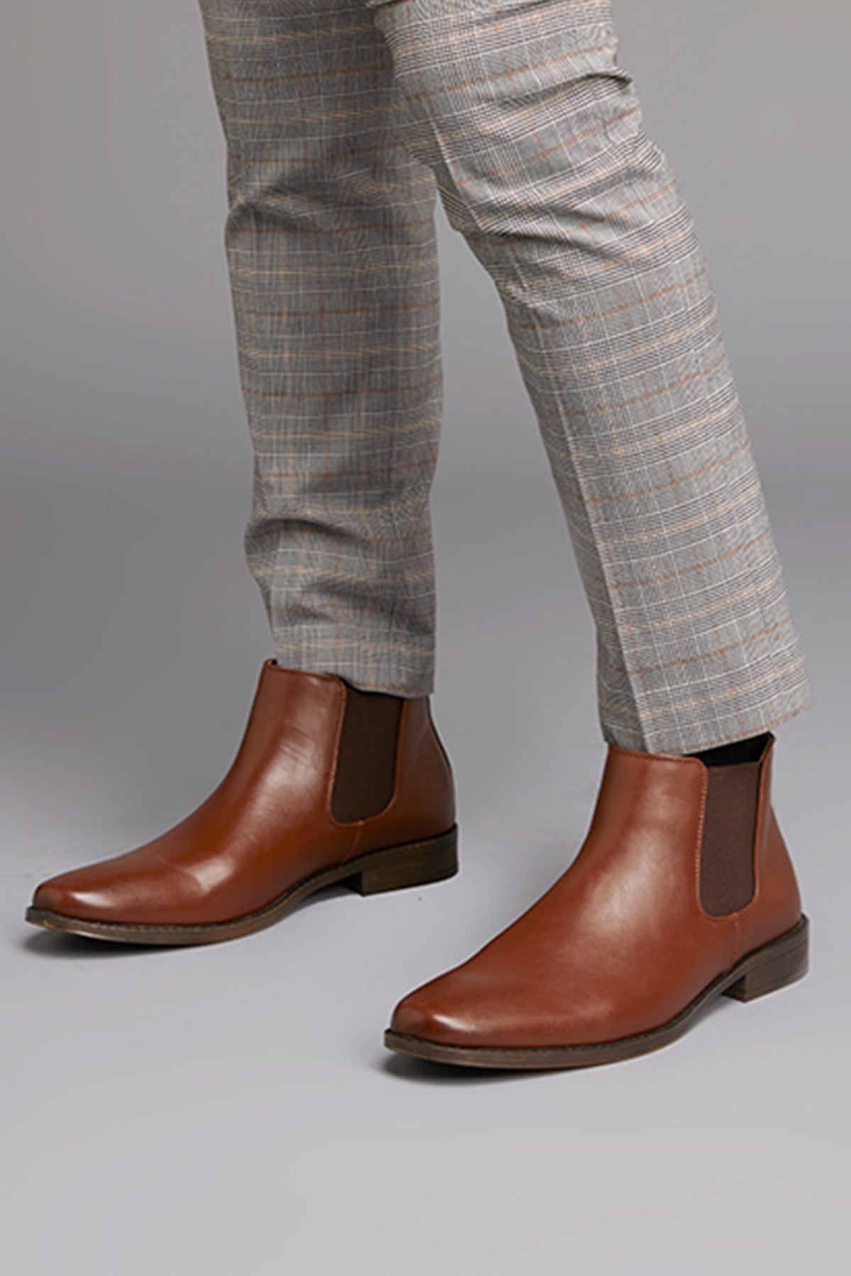 A person stands in a plain gray background, wearing UNcut Canterbury Boots in brown—a vegan Chelsea boot with elastic side panels and low heel—paired with light gray plaid pants. The design is modern yet timeless, accentuated by the footwear's sleek side gussets.