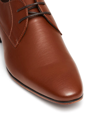 Close-up of a brown leather UNcut Chartwell Shoe with thin black laces, ideal for corporate settings or formal events. It boasts a smooth, glossy finish, slightly pointed toe, and textured upper section near the laces for contrast against the polished leather.