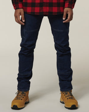 Wearing a red and black plaid shirt, tan hiking boots, and dark blue Hard Yakka Dyneema Tough Jeans, a person stands against a plain background with only their body from the shoulders down visible.