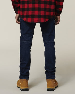 A person, facing away, wears a red and black checkered shirt, tan boots, and dark blue Hard Yakka Dyneema Tough Jeans against a plain light background.