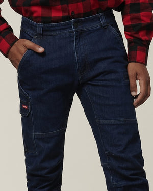 A person wears the Hard Yakka Dyneema Tough Jean, slim fit dark blue jeans made from tear and abrasion-resistant Dyneema denim, paired with a red and black plaid shirt and standing with one hand in their pocket.