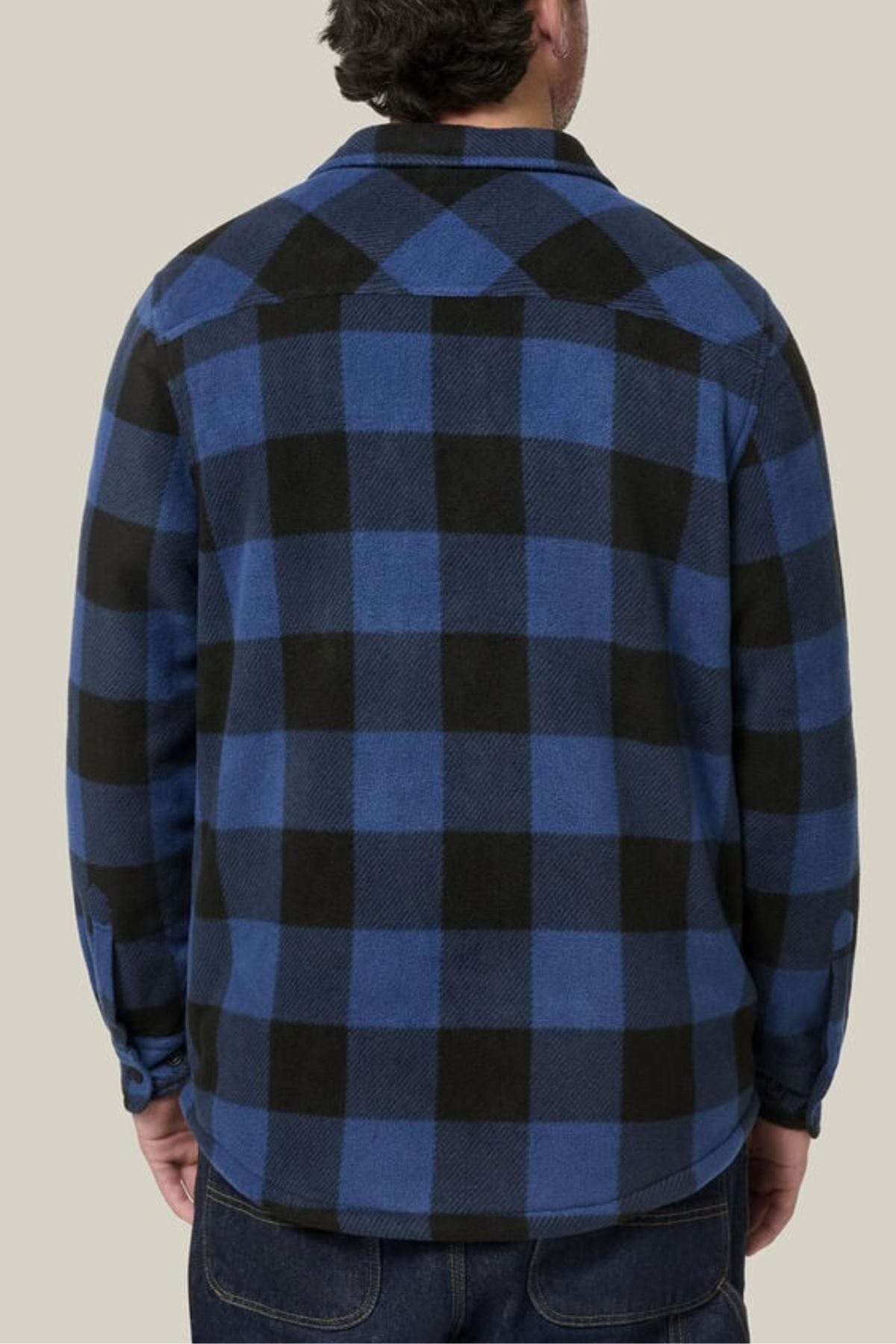 A person is seen from behind wearing the Hard Yakka Legends Sherpa with FREE Beanie—a blue and black checkered flannel shirt with fleece body lining—paired with dark jeans, standing against a plain beige background.