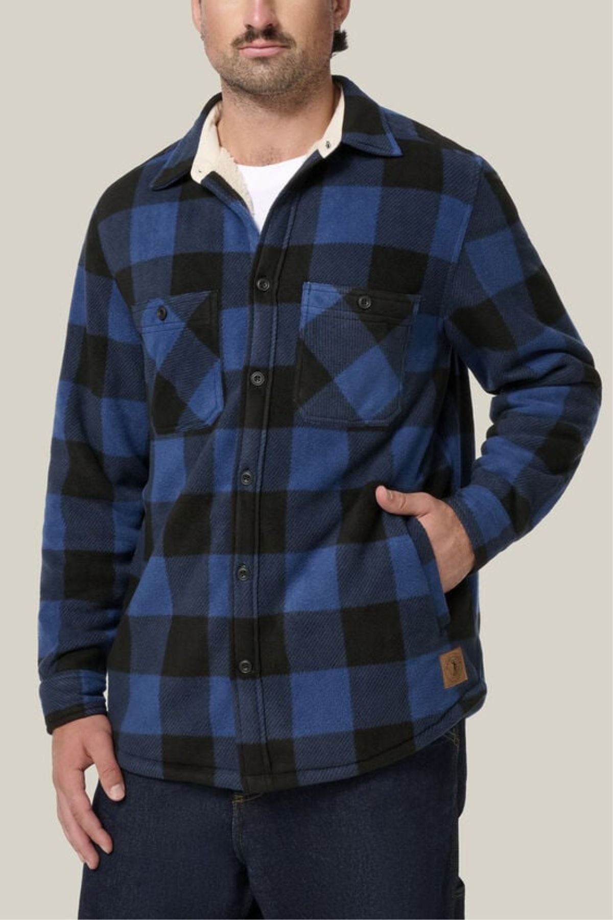 A man wears the Hard Yakka Legends Sherpa—blue and black plaid, fleece-lined, with chest pockets—and stands with one hand in his pocket against a light beige background. Includes FREE beanie.