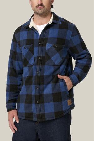 A man wears the Hard Yakka Legends Sherpa—blue and black plaid, fleece-lined, with chest pockets—and stands with one hand in his pocket against a light beige background. Includes FREE beanie.