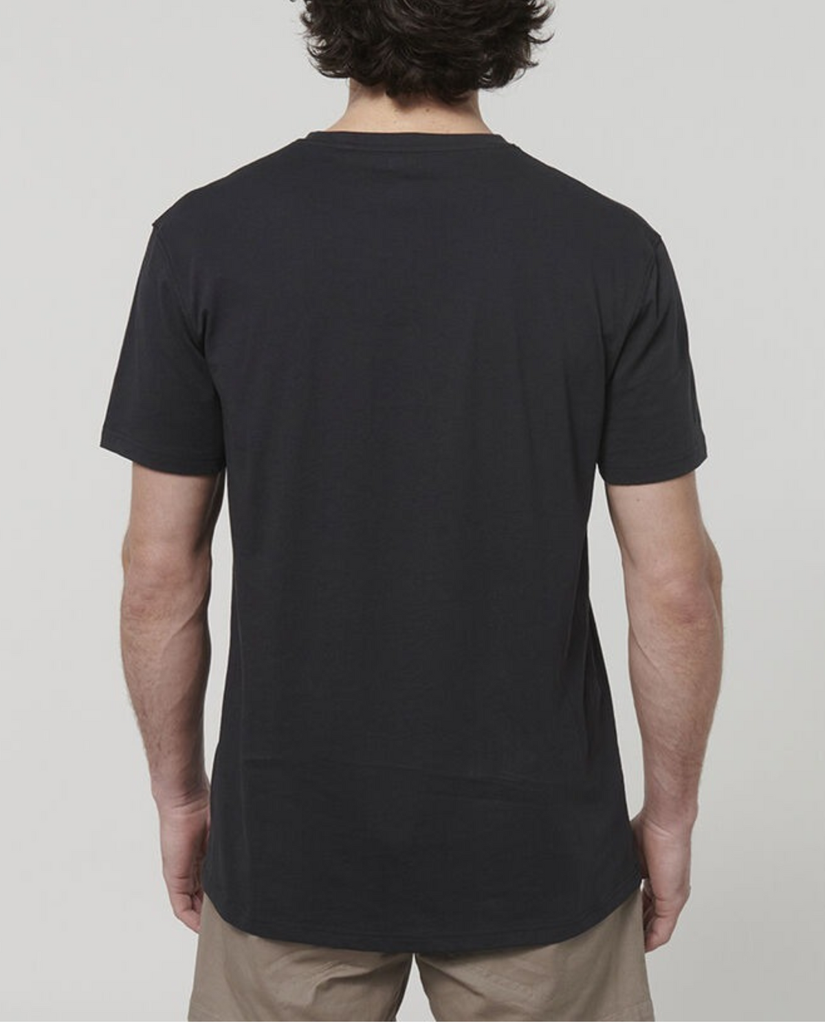 A person with short, wavy hair is seen from the back, wearing a Hard Yakka Core Tee crafted from 100% Australian cotton along with beige shorts against a plain, light grey background.