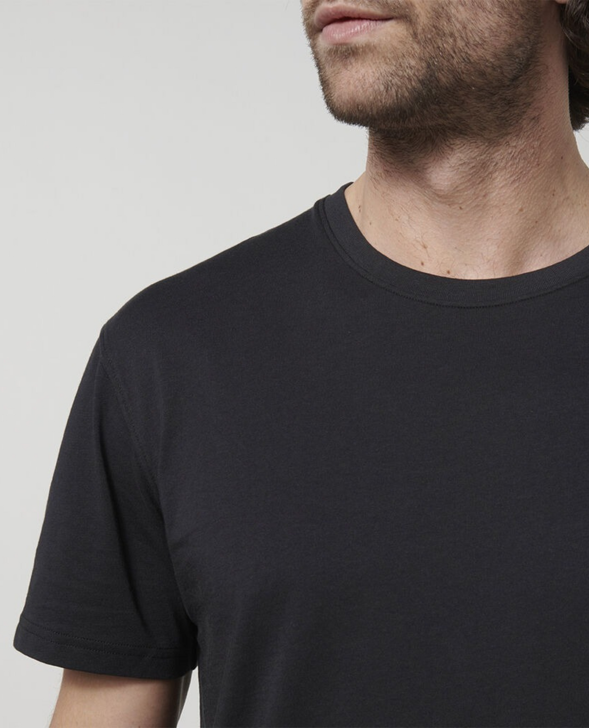 An individual with medium-length hair and a beard is seen wearing a plain black Hard Yakka Core Tee made from 100% Australian Cotton. The photo frames them from the neck down to the upper chest, highlighting the simple round neckline and short sleeves of the shirt. The background is a solid white.