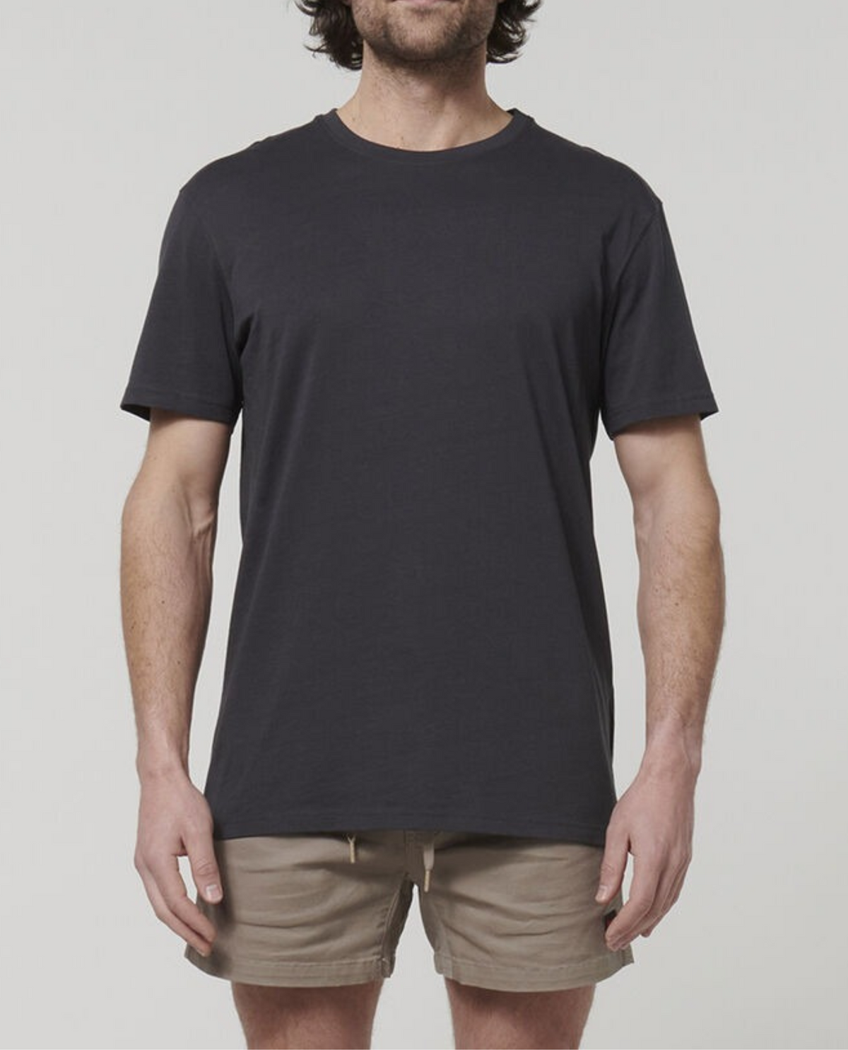 A man with medium-length hair stands in a neutral pose, wearing a plain charcoal Hard Yakka Core Tee and beige shorts made from 100% Australian Cotton. The background is a light grey color. His face is not visible, as the photo is cropped at his shoulders.
