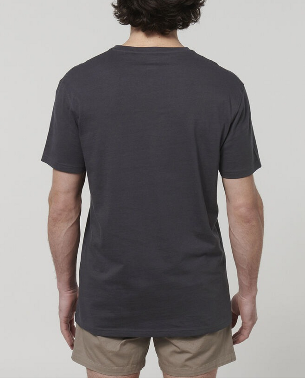 A person with short, dark hair stands with their back to the camera, dressed in a plain dark gray Hard Yakka Core Tee crafted from 100% Australian cotton and light brown shorts. The background is plain white.