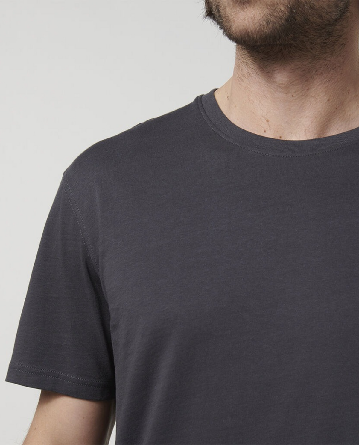 A close-up of a person wearing a dark gray Hard Yakka Core Tee, crafted from 100% cotton. The person's face is partially visible, showcasing some facial hair against a plain white background.