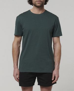 A person with medium-length dark hair is wearing a Hard Yakka Core Tee in plain dark green, made from 100% Australian Cotton, accompanied by black shorts. The individual stands against a light gray background, capturing only the upper half of their legs without including the full head.