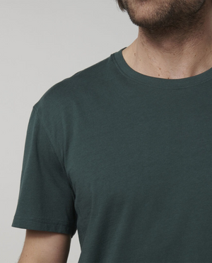 A close-up of someone wearing the Hard Yakka Core Tee, a dark green short-sleeved t-shirt crafted from 100% Australian Cotton. The image highlights the upper torso and part of the neck, with the person's face partially visible but mostly out of frame. The background is a solid light grey color, making it an ideal choice for summer work attire.
