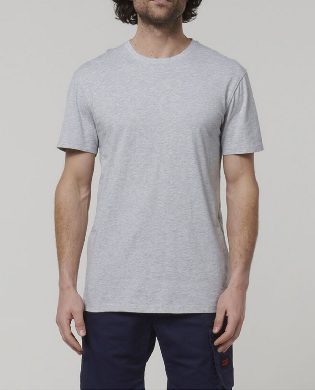 A man wearing a Hard Yakka Core Tee in a plain, light gray color, made from 100% cotton, pairs it with dark blue pants as he stands against a neutral background. His arms rest at his sides, and his head is cropped out of the image.