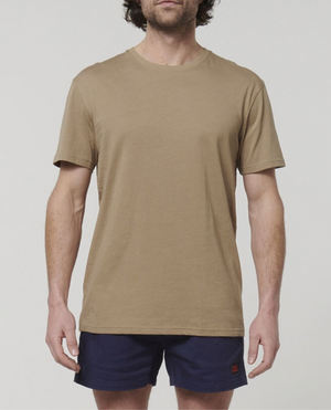 A person with medium-length dark hair is wearing a Hard Yakka Core Tee, made from 100% Australian cotton in a plain beige color, paired with dark blue shorts. They are standing facing forward against a plain white background.