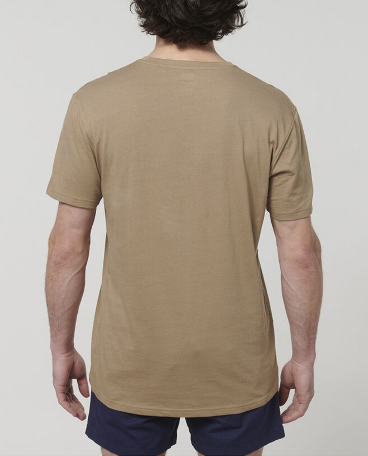 A person with short, curly hair is standing with their back to the camera. They are wearing a beige Hard Yakka Core Tee made from 100% Australian cotton and dark shorts against a plain white background.