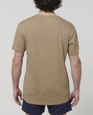 A person with short, curly hair is standing with their back to the camera. They are wearing a beige Hard Yakka Core Tee made from 100% Australian cotton and dark shorts against a plain white background.