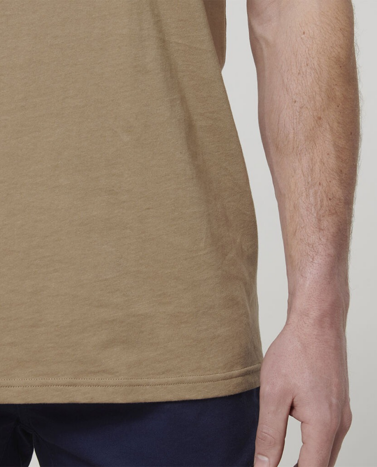 Close-up of a person's arm and hand hanging by their side, showcasing the light brown Hard Yakka Core Tee crafted from 100% Australian cotton. They are also wearing dark blue pants against a plain, light-colored background.